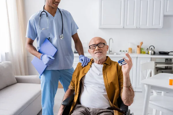 Smiling elderly man with pulse oximeter sitting in wheelchair near african american nurse with clipboard at home — Fotografia de Stock