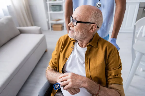 Smiling senior man with pulse oximeter looking away near african american nurse at home — Stockfoto