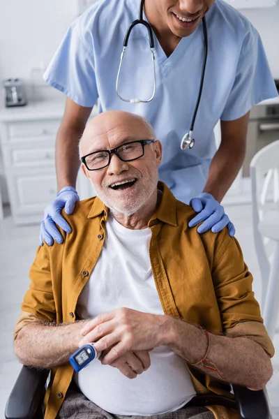 Cheerful elderly man with pulse oximeter looking at camera near african american nurse at home - foto de stock