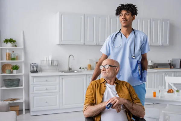Positive african american nurse holding clipboard near pensioner with pulse oximeter at home — Stockfoto