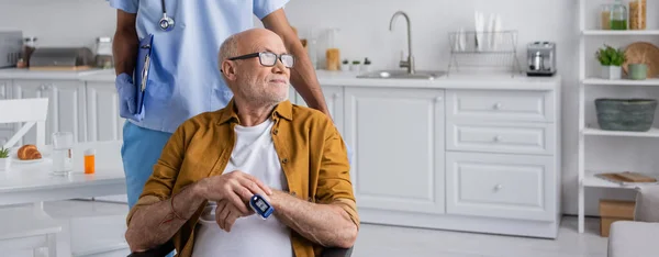 Старший чоловік з імпульсним оксиметром, який дивиться далеко від афроамериканської медсестри з буфетом вдома, банер — стокове фото