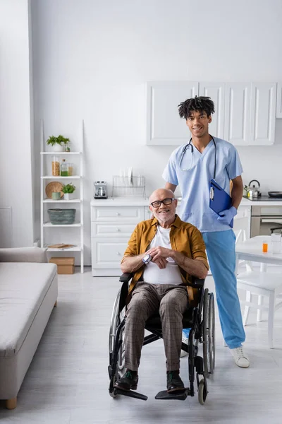 Smiling african american nurse with clipboard looking at camera near pensioner with pulse oximeter in wheelchair at home - foto de stock