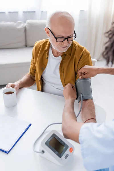 African american nurse fastening tonometer on hand of senior patient near tea and clipboard at home — Stockfoto