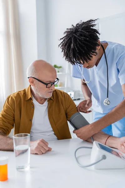 African american nurse fastening tonometer on hand of elderly patient near water and pills at home - foto de stock