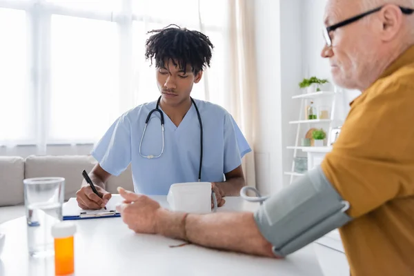 African american nurse writing on clipboard near tonometer and blurred pensioner at home — Foto stock