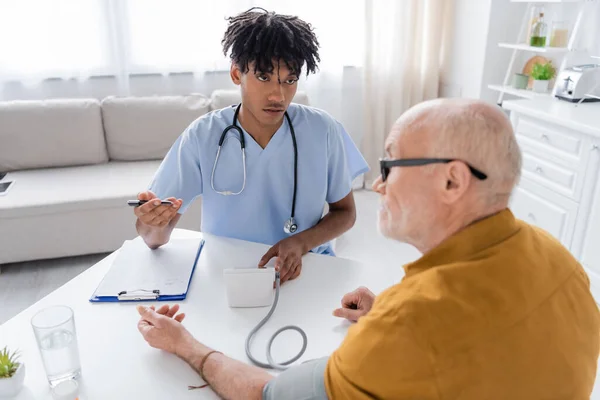 African american nurse talking to senior patient near tonometer and clipboard at home — Foto stock