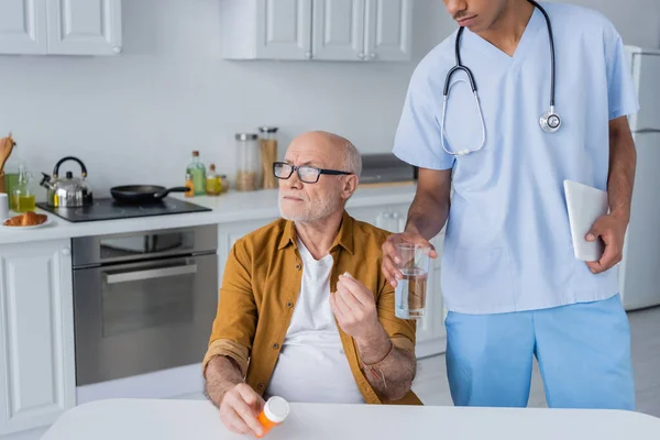 Elderly man holding pills near african american nurse with digital tablet and glass of water at home — Fotografia de Stock