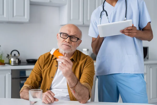 Senior man holding pills near water and african american nurse using digital tablet in kitchen — Stock Photo