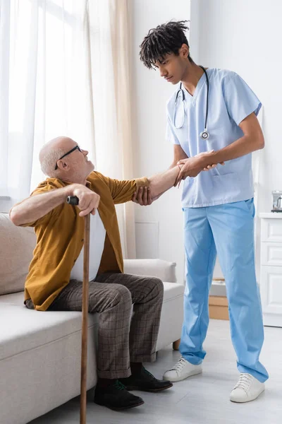 African american nurse helping pensioner with walking cane on couch at home — Stock Photo