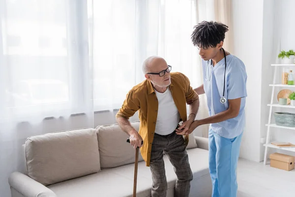 African american nurse in uniform helping elderly man with walking cane at home — Foto stock