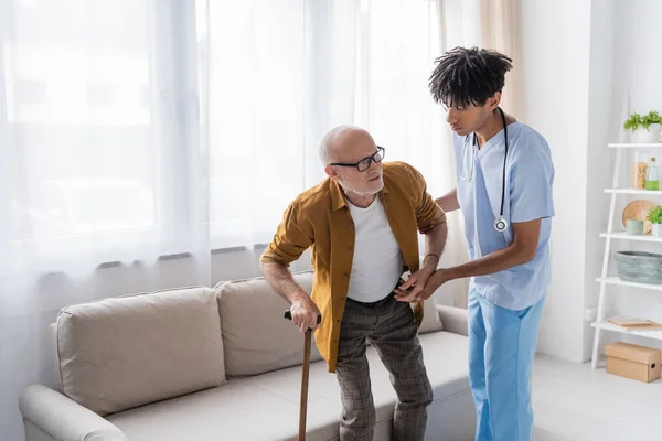 African american nurse in uniform helping elderly man with walking cane at home - foto de stock