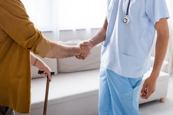 Cropped view of senior man with walking cane shaking hand of african american nurse at home — Fotografia de Stock