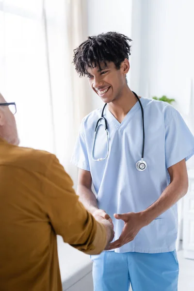 Smiling african american nurse shaking hand of elderly patient at home — Foto stock