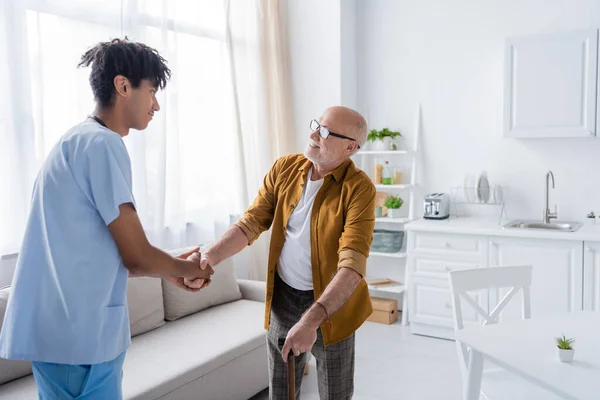 Smiling elderly patient with walking cane shaking hand of african american nurse at home - foto de stock