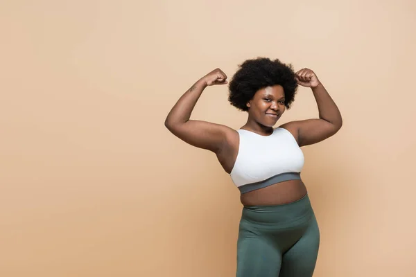 Cheerful african american plus size woman in crop top showing power gesture isolated on beige — Stock Photo