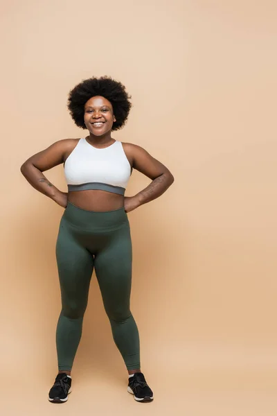 Full length of happy african american plus size woman in crop top and leggings posing with hands on hips on beige — Stock Photo