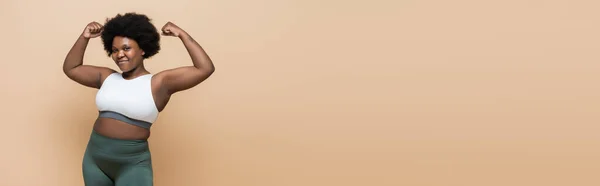 Cheerful african american plus size woman in crop top showing power gesture isolated on beige, banner — Stock Photo