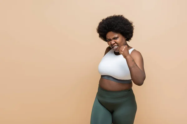 Emotional african american plus size woman in crop top showing clenched fist isolated on beige - foto de stock