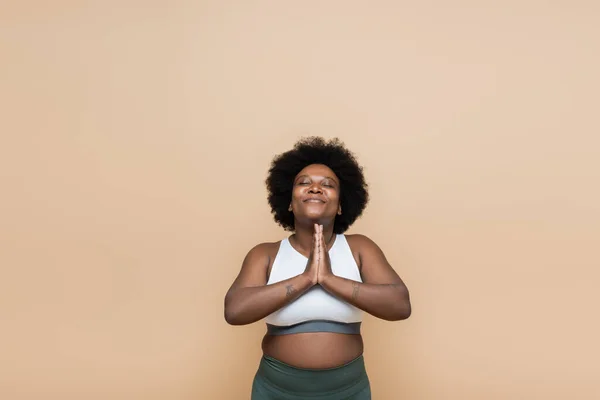 Pleased african american plus size woman in crop top praying isolated on beige — Stock Photo
