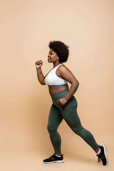 Full length of happy african american plus size woman in sportswear running on beige — Photo de stock