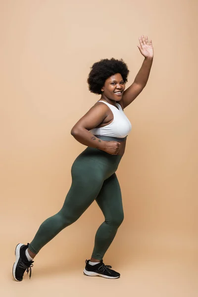 Повна довжина щасливої афроамериканки плюс розмір жінки в спортивному одязі біжить і махає рукою на бежевому — стокове фото
