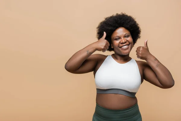 Happy african american plus size woman in crop top showing thumbs up isolated on beige — Foto stock