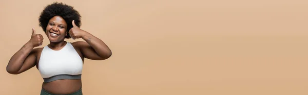 Happy african american plus size woman in crop top showing thumbs up isolated on beige, banner — Stock Photo