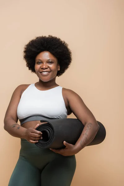 Joyful african american plus size woman in crop top holding fitness mat isolated on beige — Stock Photo