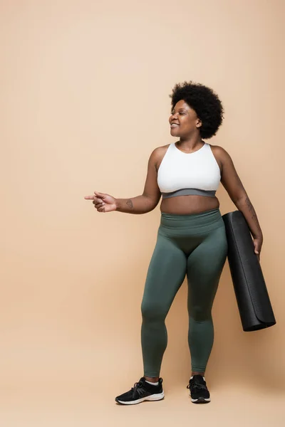 Full length of happy african american plus size woman holding fitness mat and pointing with finger on beige — Fotografia de Stock