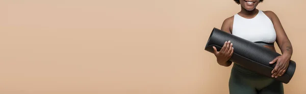 Cropped view of happy african american plus size woman with tattoo holding fitness mat isolated on beige, banner — Stock Photo