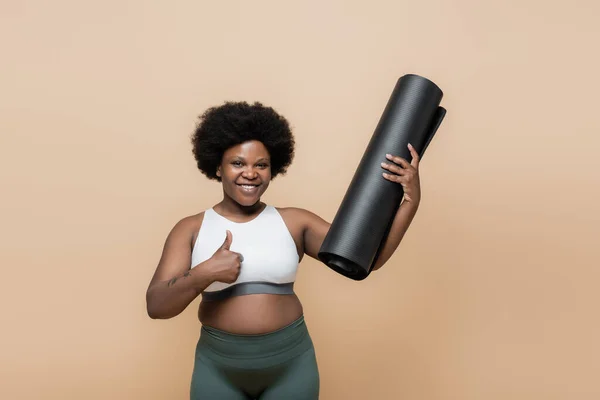 Smiling african american plus size woman in crop top holding fitness mat and showing thumb up isolated on beige - foto de stock