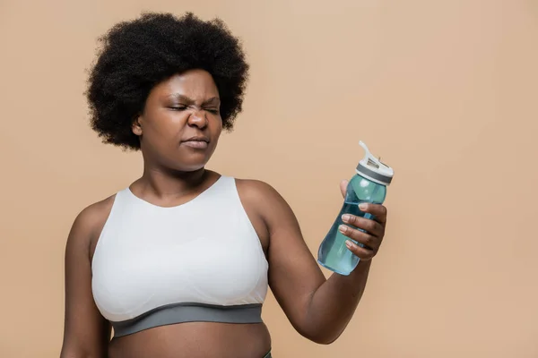 Displeased african american plus size woman in crop top holding sports bottle with water isolated on beige — Stockfoto