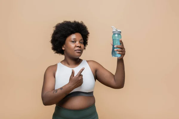 African american plus size woman in crop top holding sports bottle with water and pointing at herself isolated on beige - foto de stock