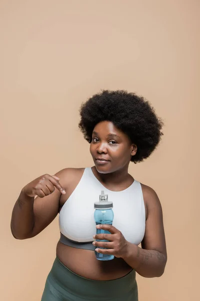 African american plus size woman in crop top pointing at sports bottle with water isolated on beige — Foto stock