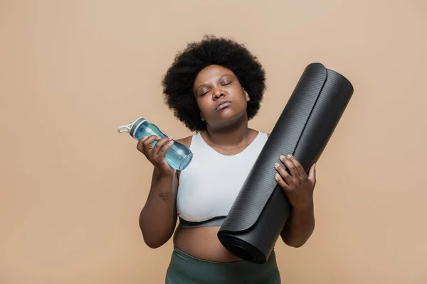 Curly african american plus size woman with closed eyes holding fitness mat and sports bottle isolated on beige — Stock Photo
