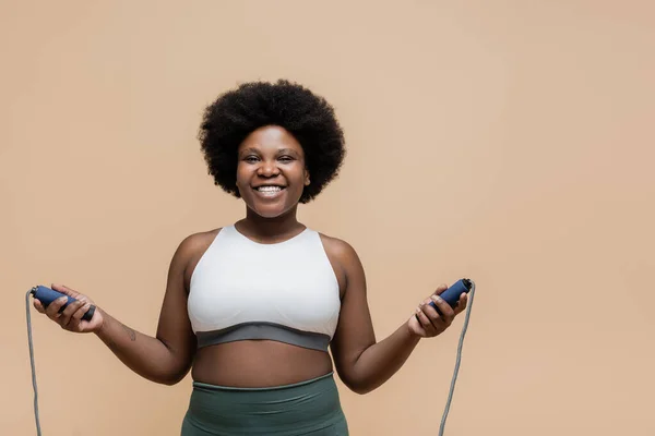 Smiling african american plus size woman in sportswear holding skipping rope isolated on beige — Stockfoto