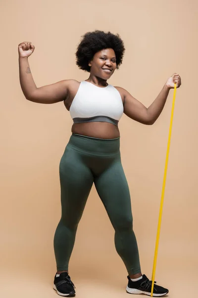Full length of joyful african american plus size woman in sportwear exercising with resistance band on beige — Stock Photo
