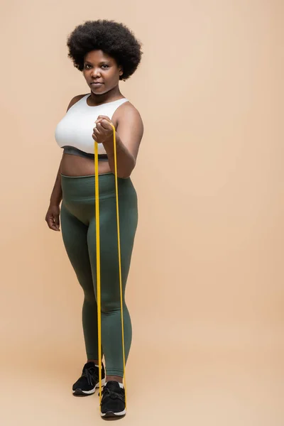 Full length of african american plus size woman in sportwear exercising with resistance band on beige — Stock Photo