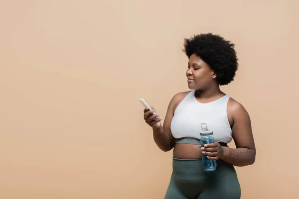 Curly african american plus size woman holding sports bottle with water and using smartphone isolated on beige — Stock Photo