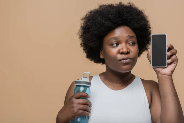 Curious african american plus size woman holding sports bottle and smartphone with blank screen isolated on beige — Photo de stock