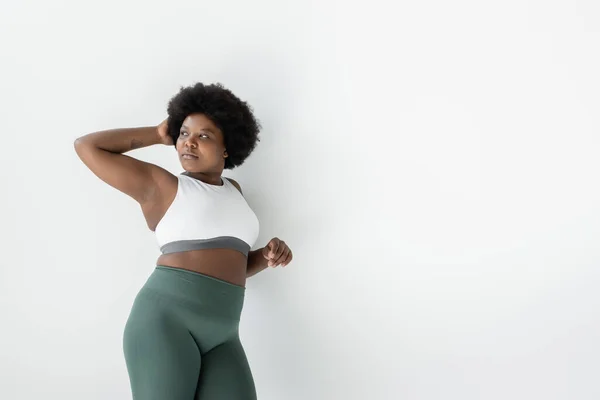 Curly african american plus size woman in sportswear standing near white wall — Stock Photo