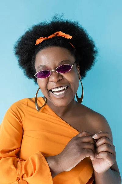 Positive african american plus size woman in sunglasses and hoop earrings isolated on blue — Stock Photo