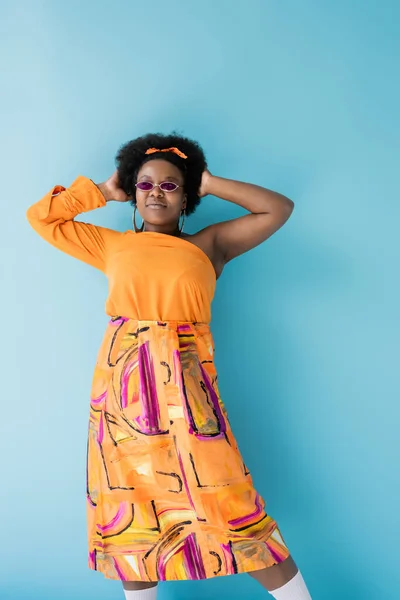 Curly african american plus size woman in sunglasses and orange dress posing on blue — Fotografia de Stock