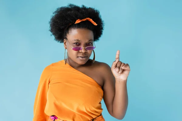 Curly african american plus size woman in sunglasses and hoop earrings pointing up isolated on blue — Stock Photo