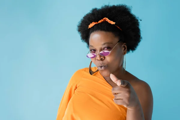 Curly african american woman in pink sunglasses pointing with finger isolated on blue — Stockfoto