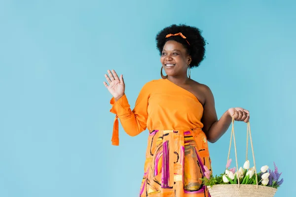 Cheerful african american body positive woman holding straw bag with flowers isolated on blue — Photo de stock