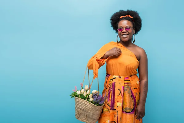 Жизнерадостная африканская американское тело позитивная женщина в солнечных очках, держа соломенный мешок с цветами изолированы на синий — стоковое фото