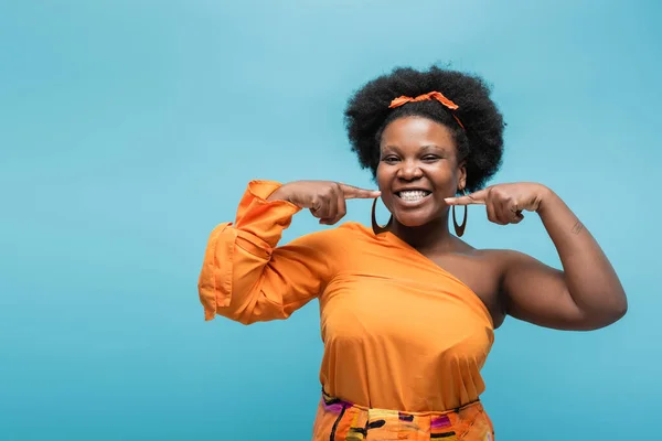 Cheerful african american body positive woman in orange dress and hoop earrings pointing at cheeks isolated on blue - foto de stock