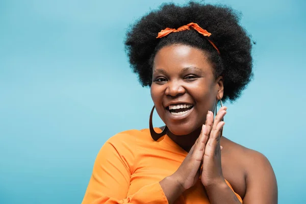 Happy african american body positive woman in orange dress and hoop earrings isolated on blue — Foto stock