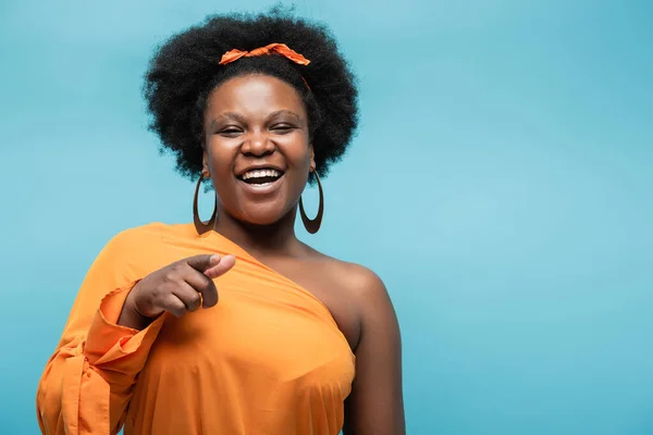 Smiling african american body positive woman in orange dress and hoop earrings pointing with finger isolated on blue — Foto stock